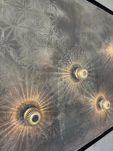 Wand mit dezenten Mustern und mehreren strahlenden Deckenleuchten.